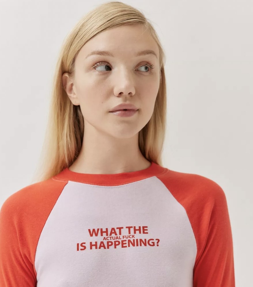A person with long blonde hair wearing a white and red baseball tee that says "what the actual fuck is happening?" on it