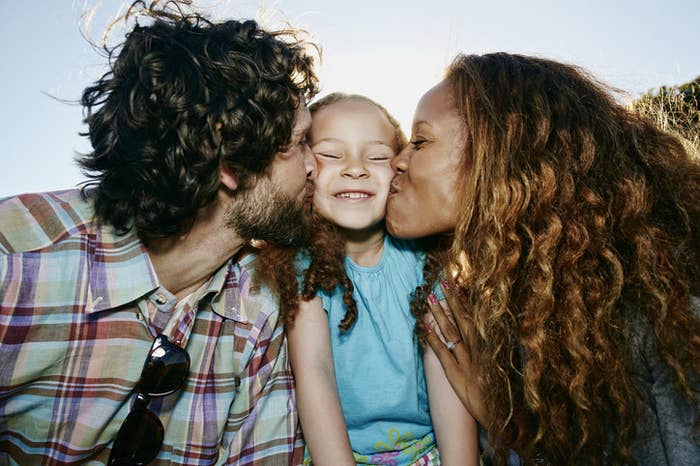 A white man and a Black woman lean over to kiss the light-skinned girl between them, who is smiling with her eyes closed