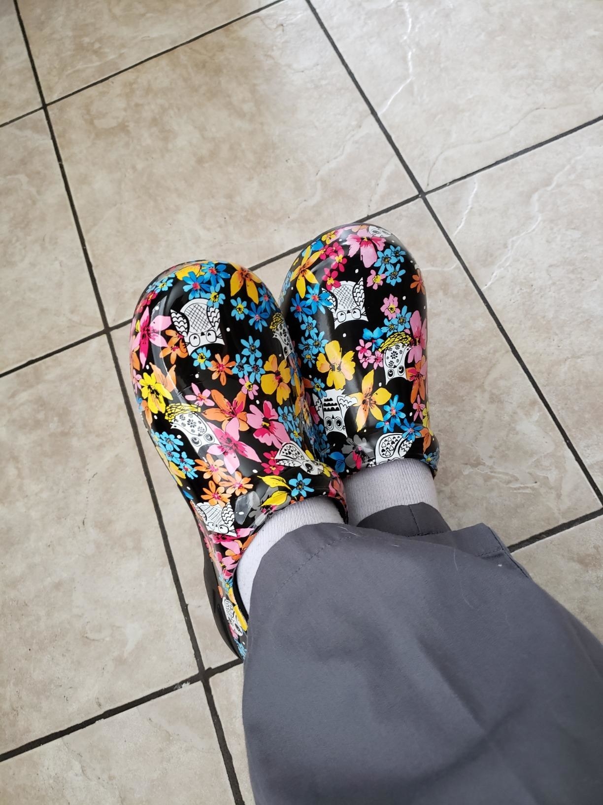 Reviewer in scrubs with ankles crossed wearing the black with colorful floral design clogs