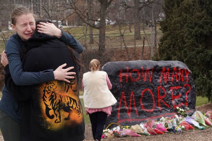 Two people hold each other and cry near a boulder, which has bouquets of flowers at its base, spray-painted with the message "how many more?"