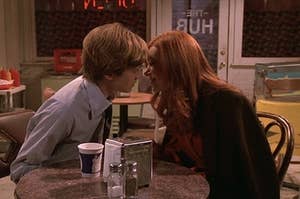 Eric and Donna sit at a table in The Hub and lean in to kiss each other