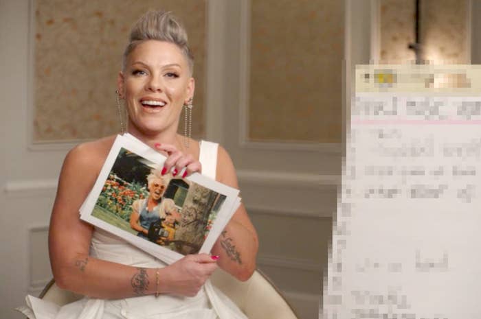 Pink sits in a room and holds up a still from her so what music video and smiles and white board beside her is pixelated