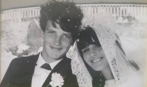 A smiling bride and groom, wearing wedding attire, sit close together under a canopy