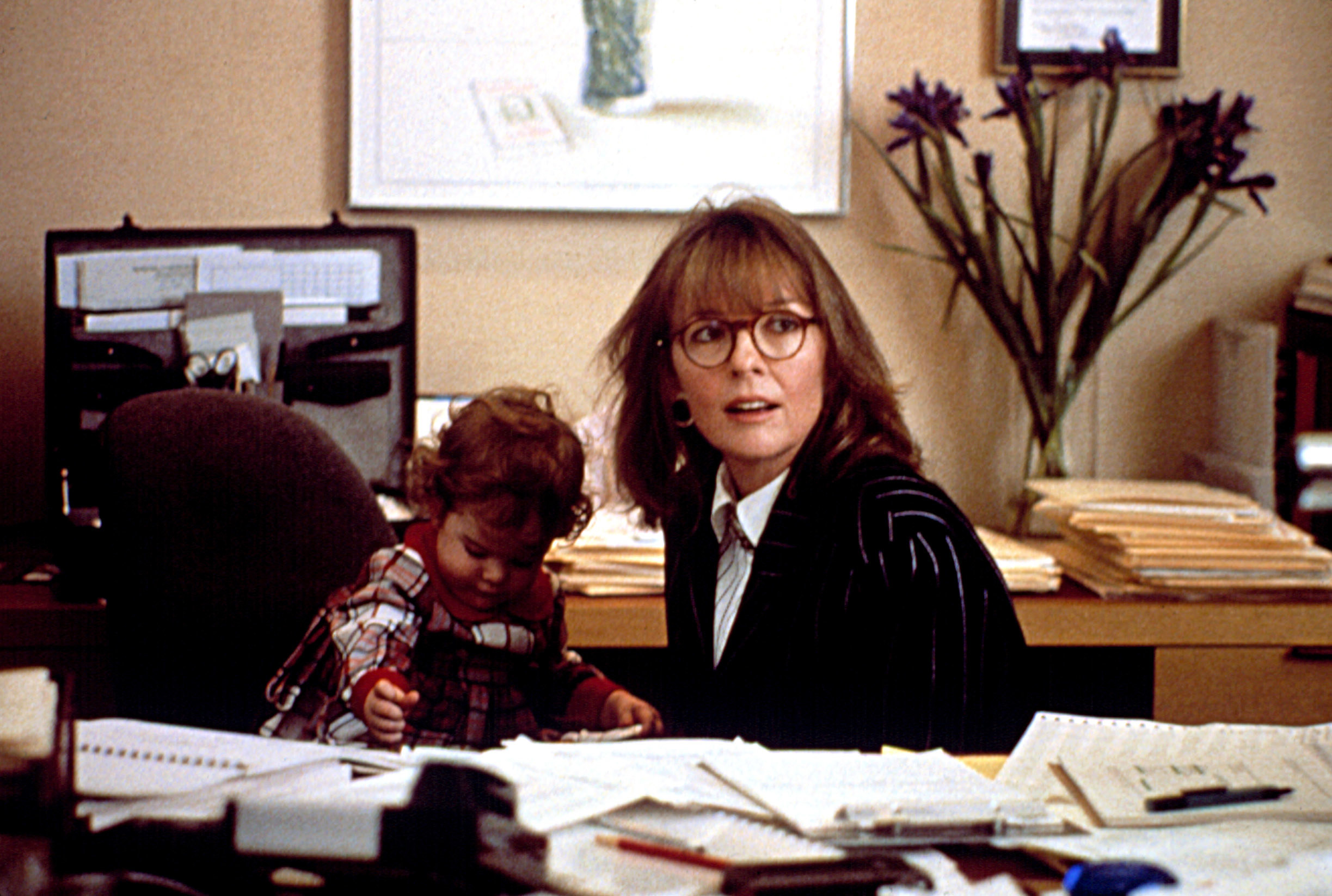 Diane Keaton sits at a desk covered in papers as J.C. while a baby sits in a chair next to her