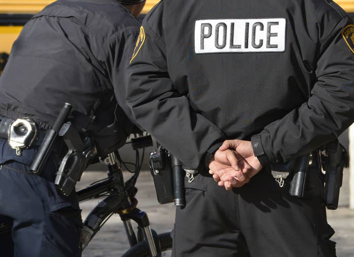 Two officers stand side by side from the neck down, one wears a jacket that says &quot;police&quot; and has their hands folded behind their back; their guns, handcuffs, and utility belts are visible