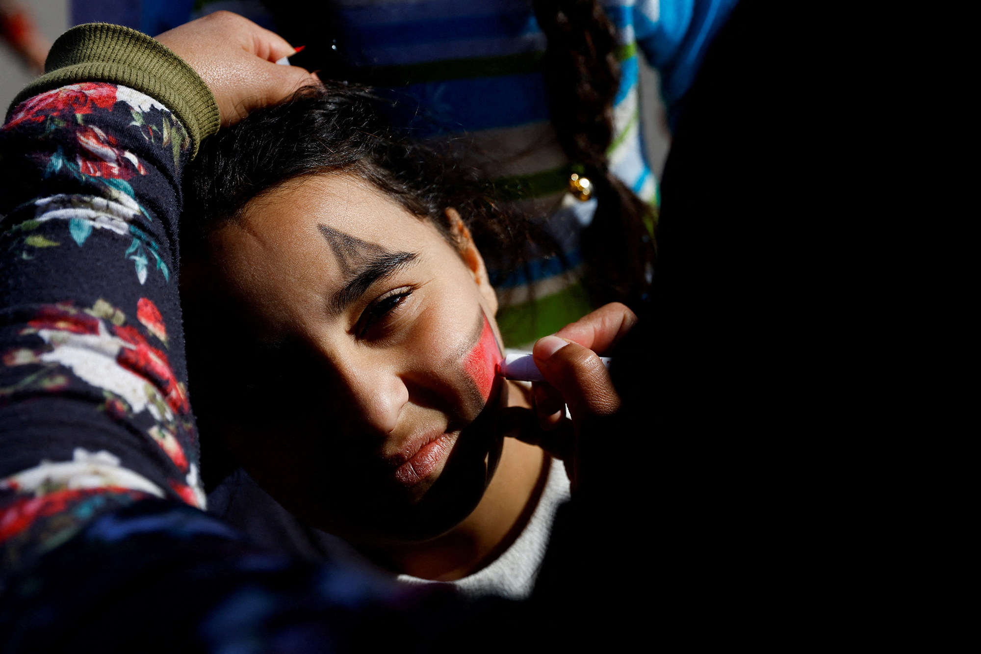 a serious looking child gets their face painted by someone who is hidden in the shadows