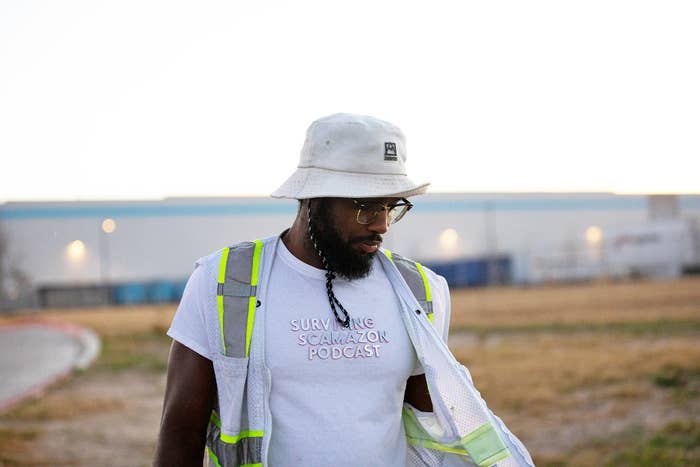 a man in a bucket hat and neon vest stands in front of a huge amazon warehouse