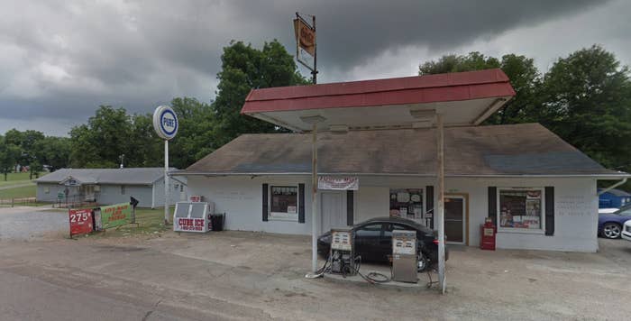 A black sedan is parked behind two gas pumps and under a red awning at a rural gas station