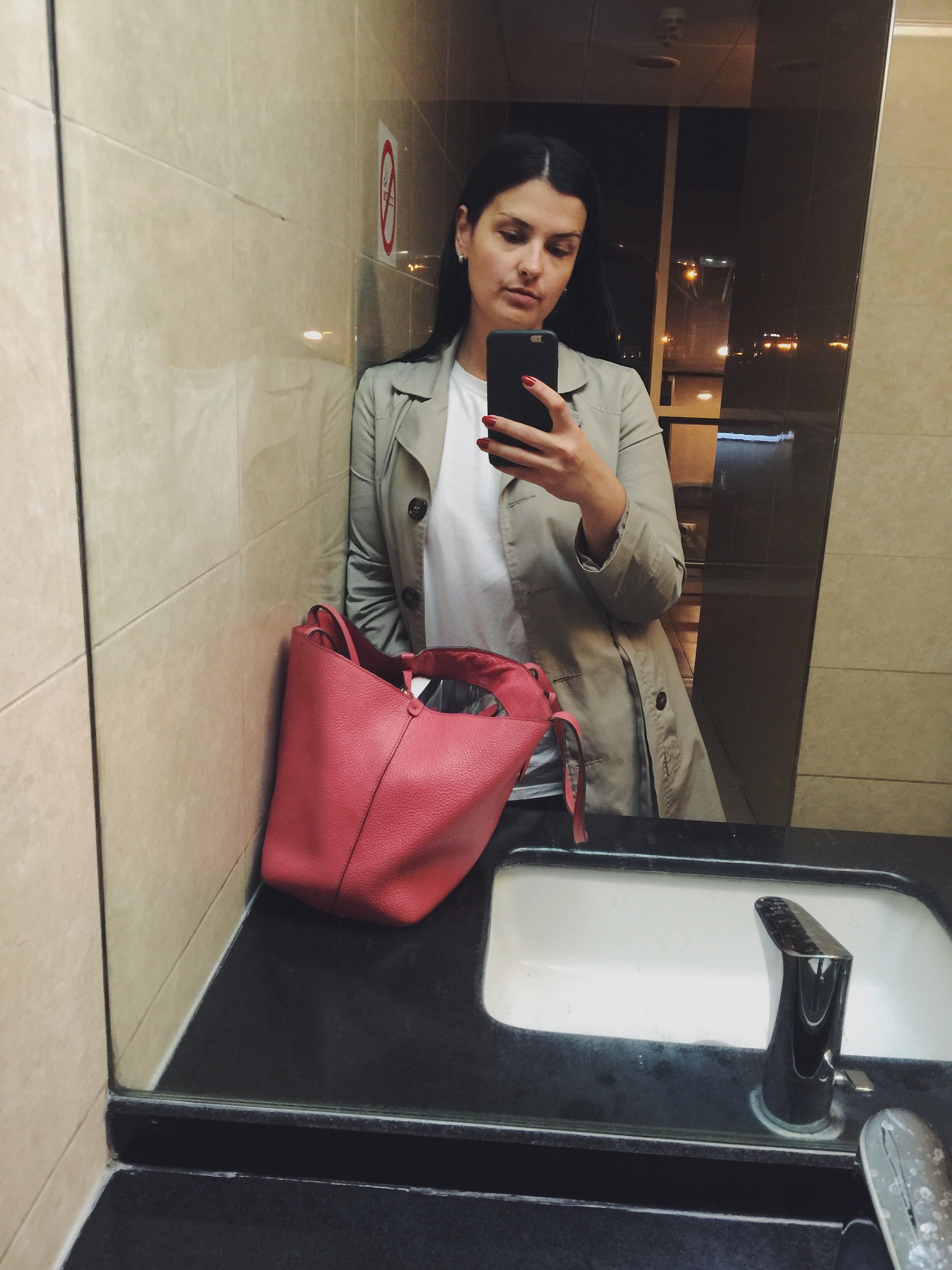 A woman taking a selfie in front of a bathroom window, with her large bag on the sink countertop