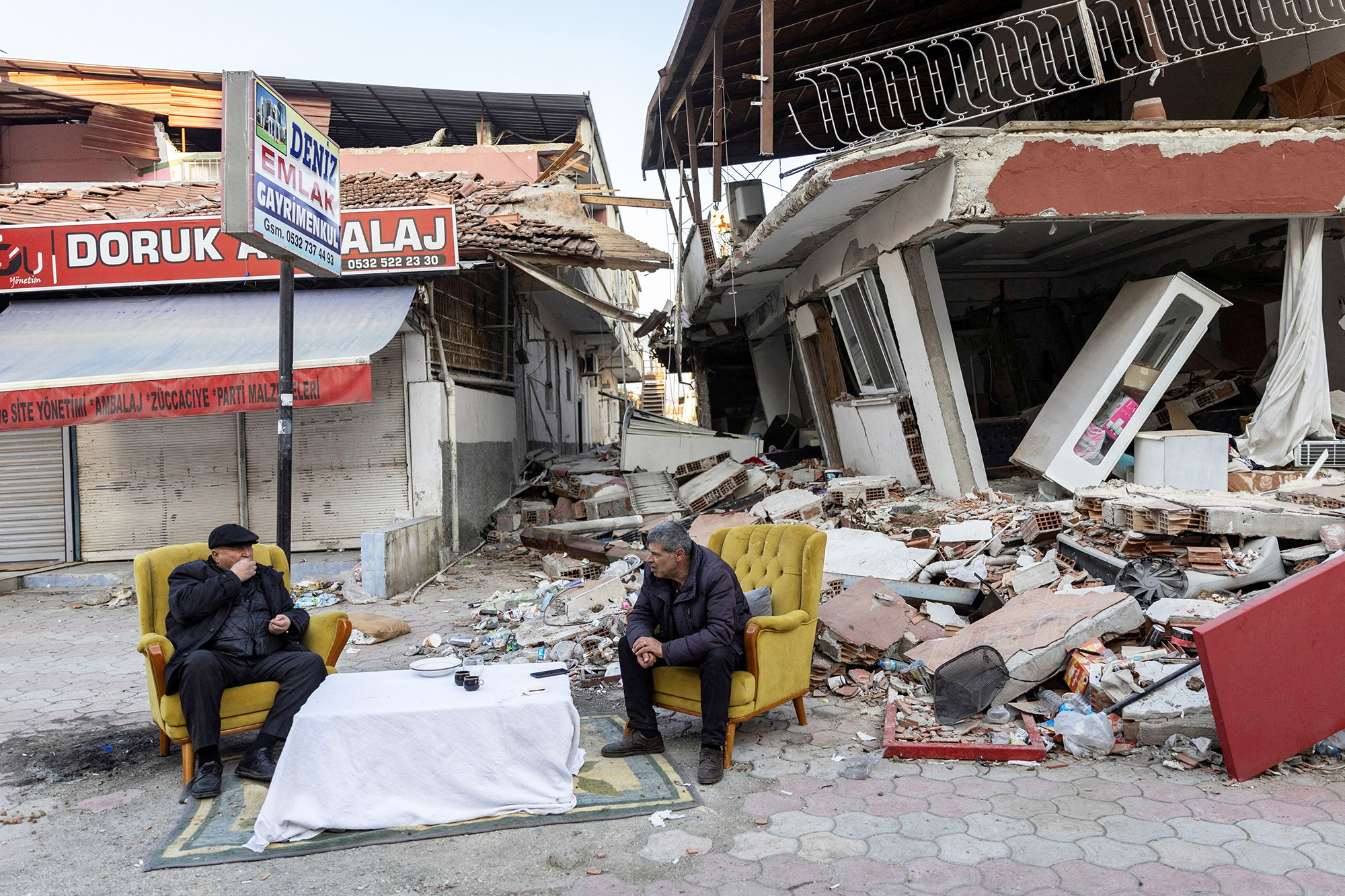 two men sit by a makeshift table in front of a collapsed building