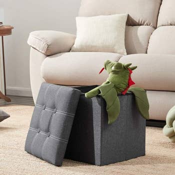 the grey cube-shaped storage ottoman with the lid off and objects stored inside