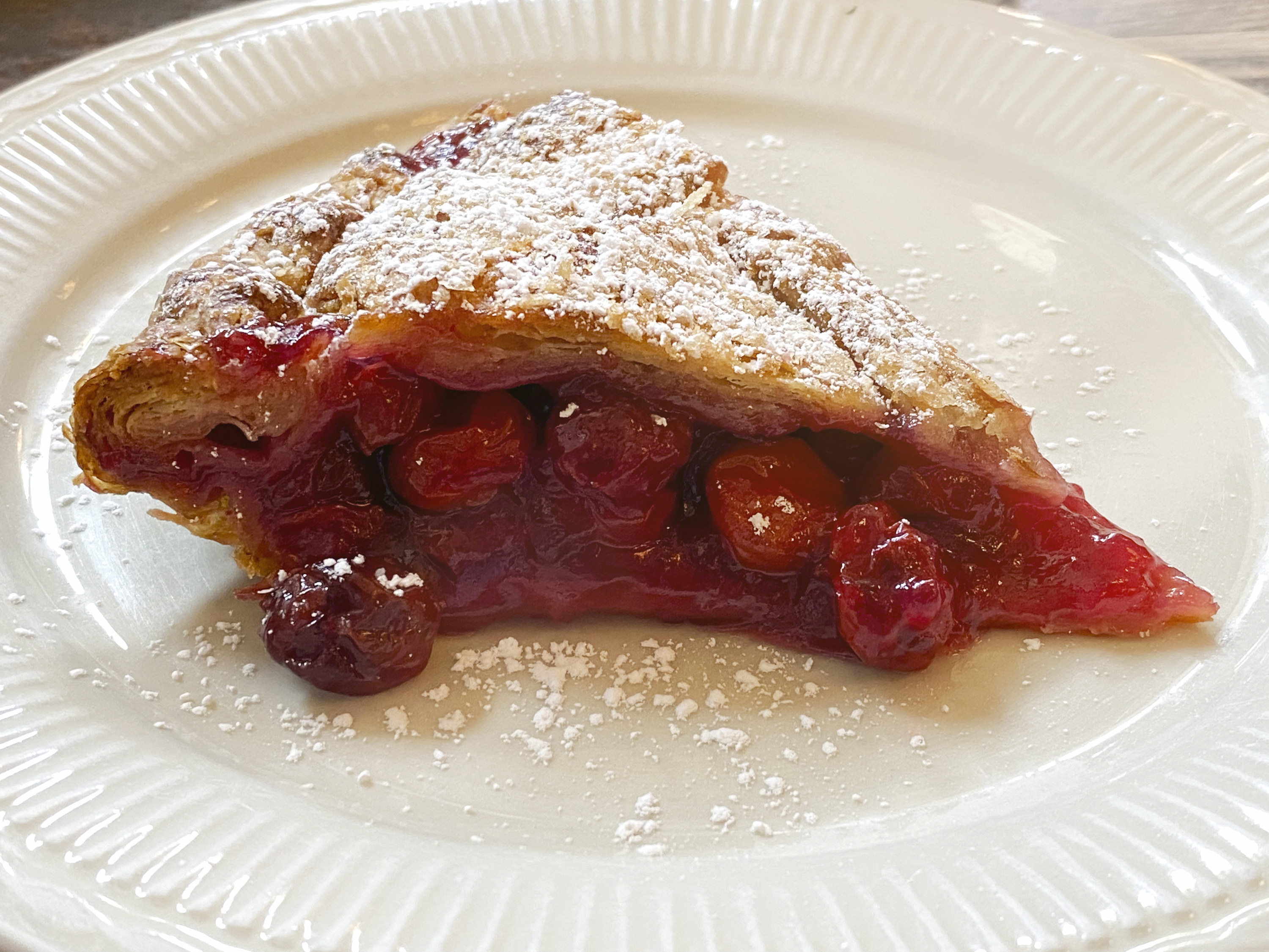 An image of a slice of cherry pie on a white plate, with icing sugar dusted on top of the pie&#x27;s crust