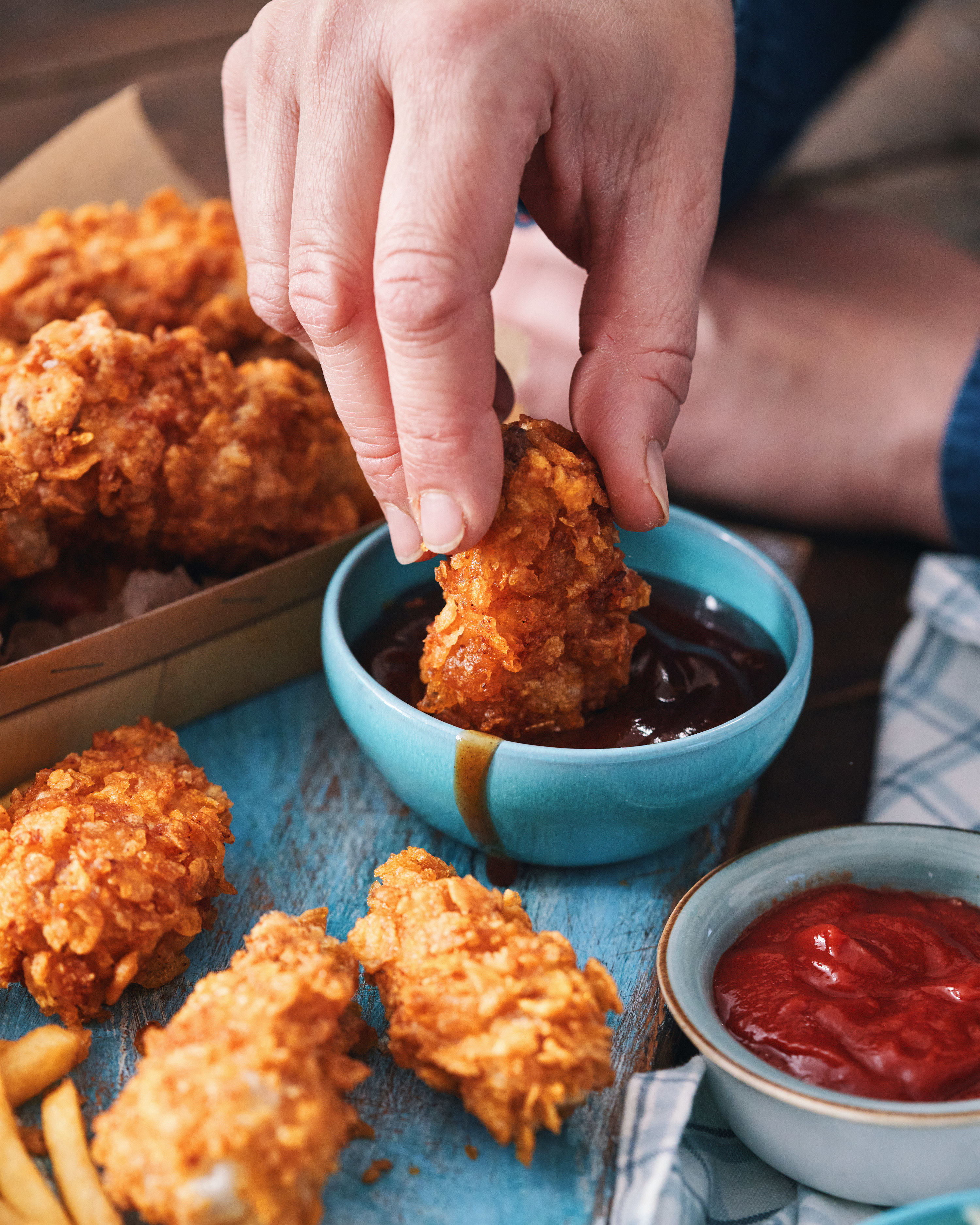 An image of a person&#x27;s hand dipped a piece of fried chicken into a small blue bowl of barbeque sauce.