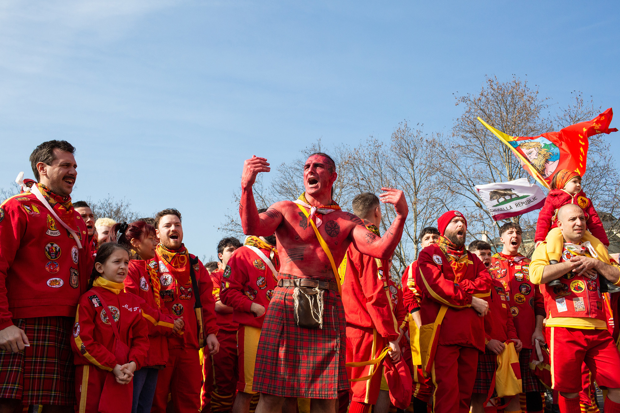 People wear red outfits and yellow bandanas outside, all yelling, surrounding a shirtless man wearing a kilt whose torso is painted red