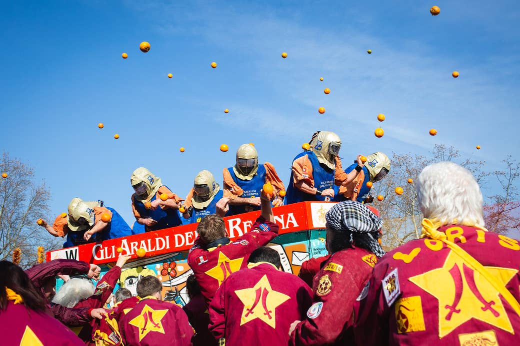 People wearing jackets depicting two swords inside a yellow heart toss oranges at a group of people wearing helmets, vests. The bright blue sky is punctuated with dozens of oranges flying midair