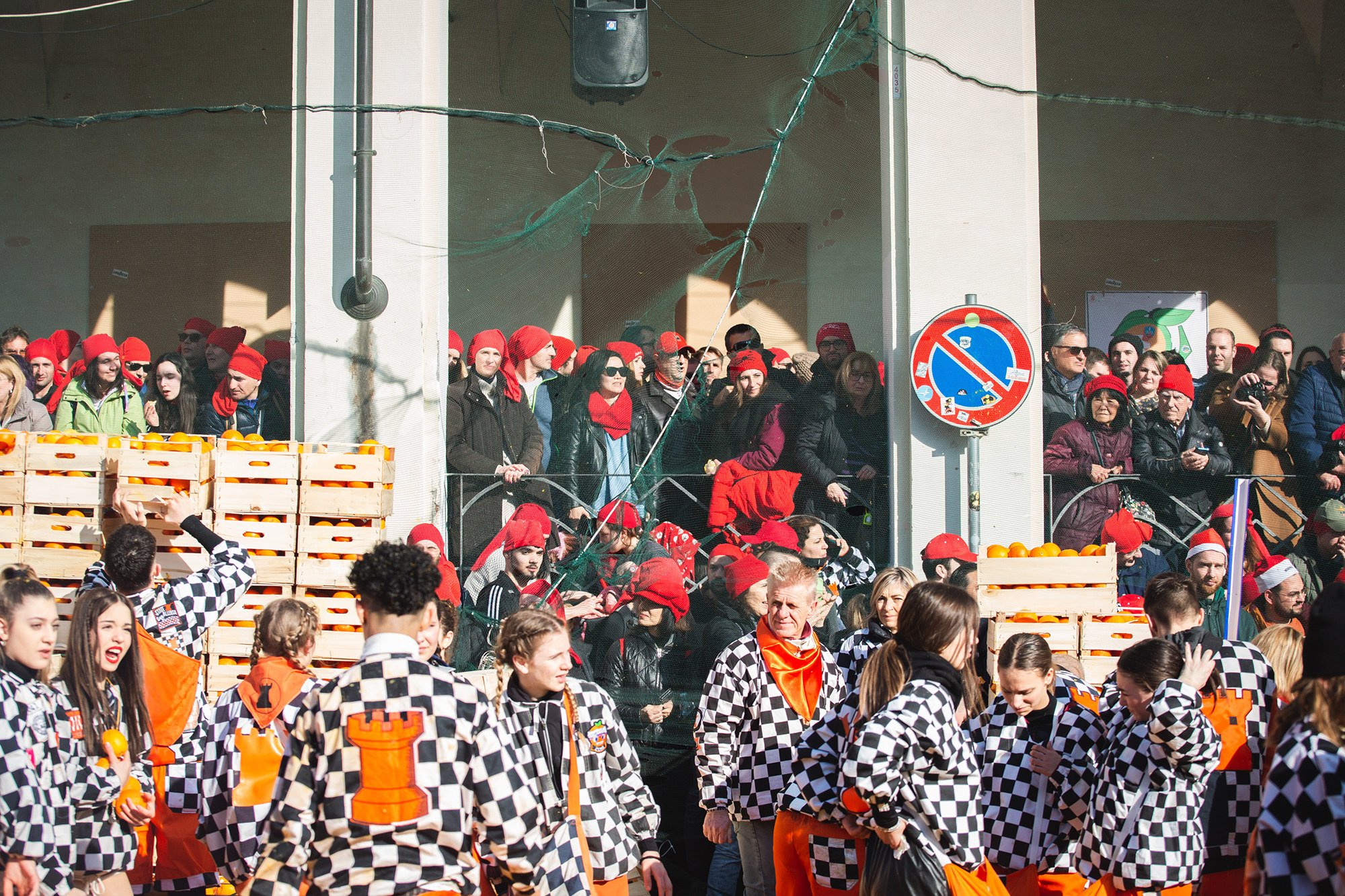 People wear checkered shirts with the rook chess piece on the backs, standing near crates of oranges while people in the background wear red beanies, standing behind a very tall net