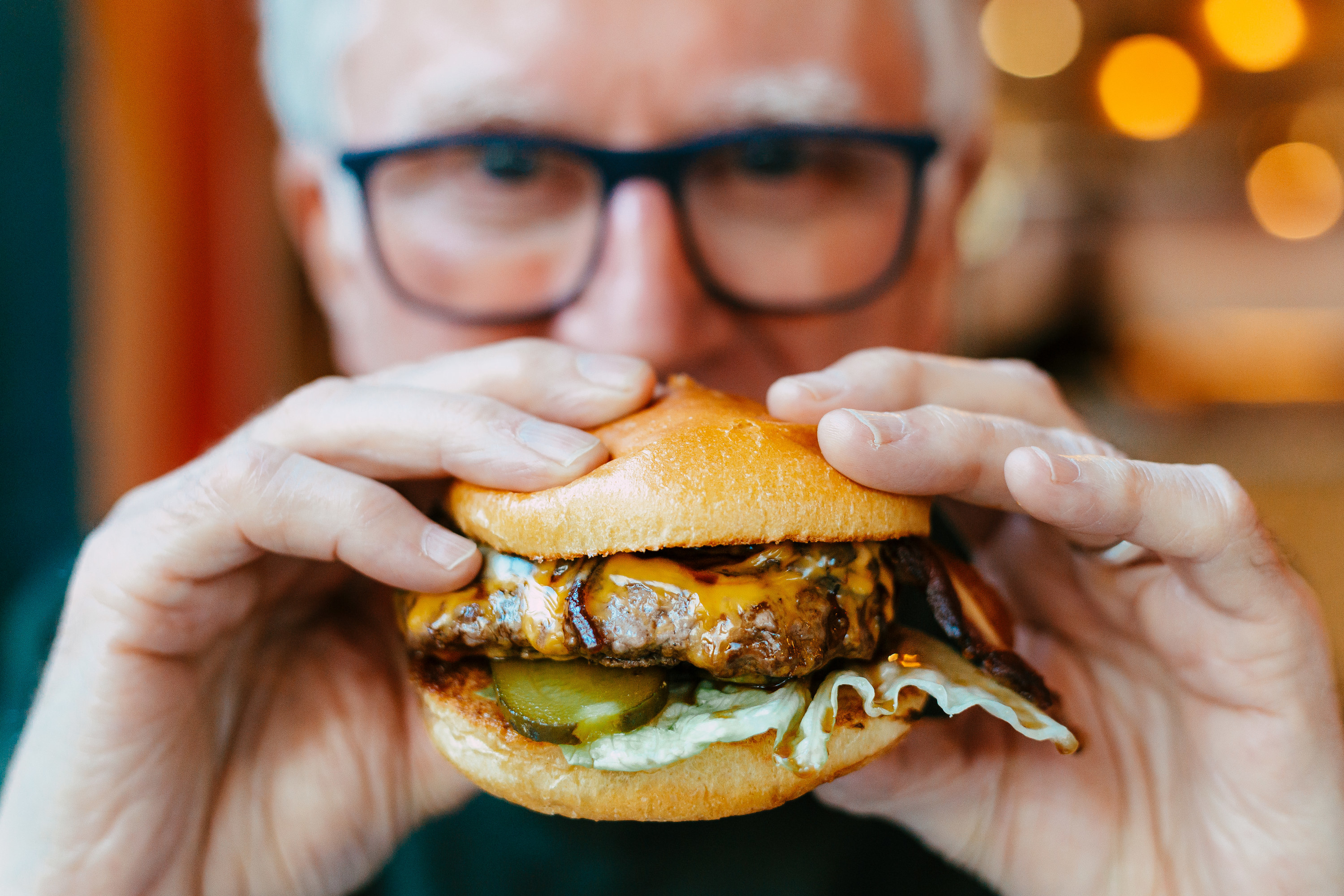An image of an older man wearing glasses, holding a burger in front of him