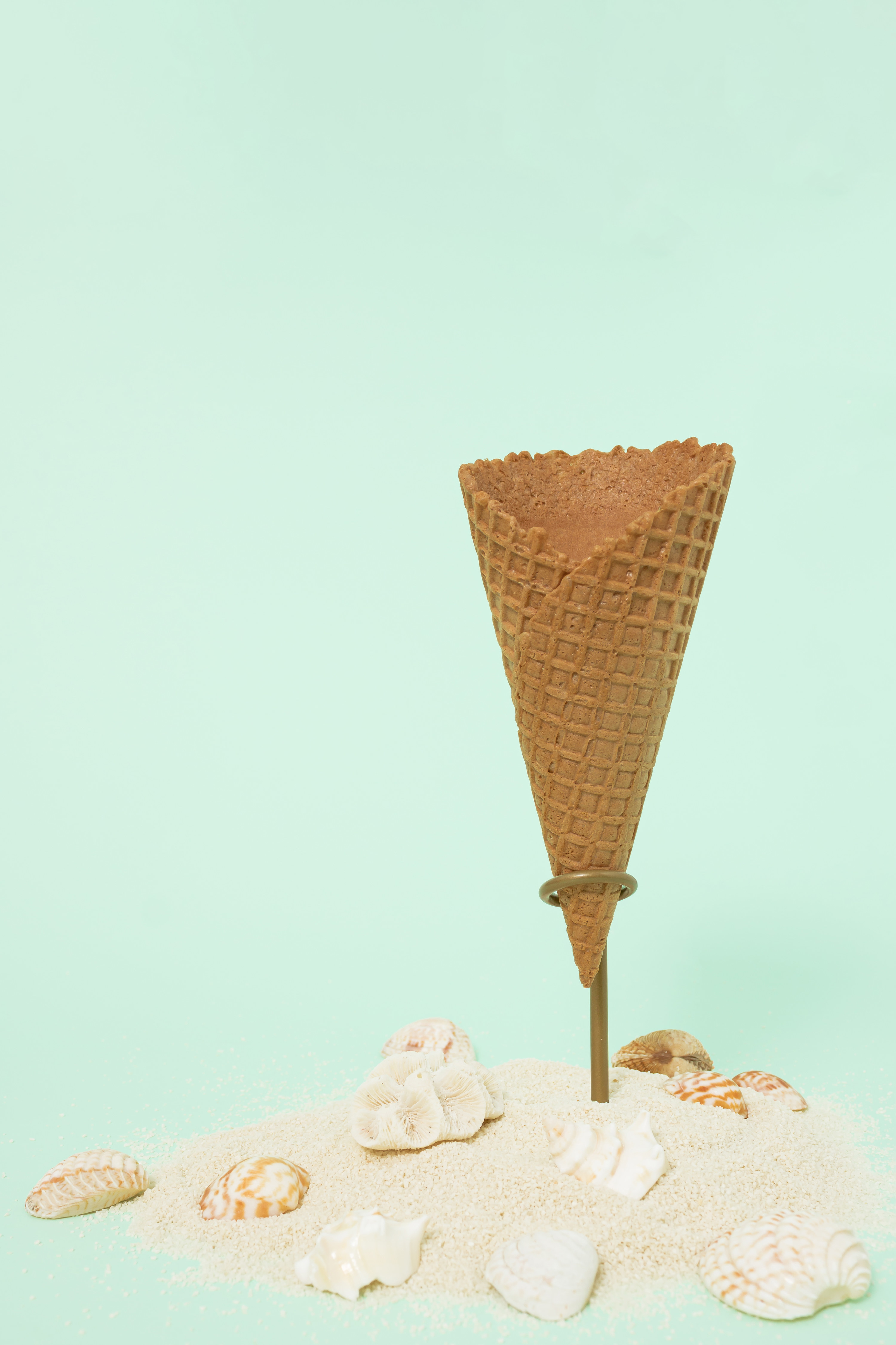 An image of a single ice cream cone without the ice cream, in a cone stand against a mint green background. There are shells and a sprinkling of sand on the ground in front of the cone.