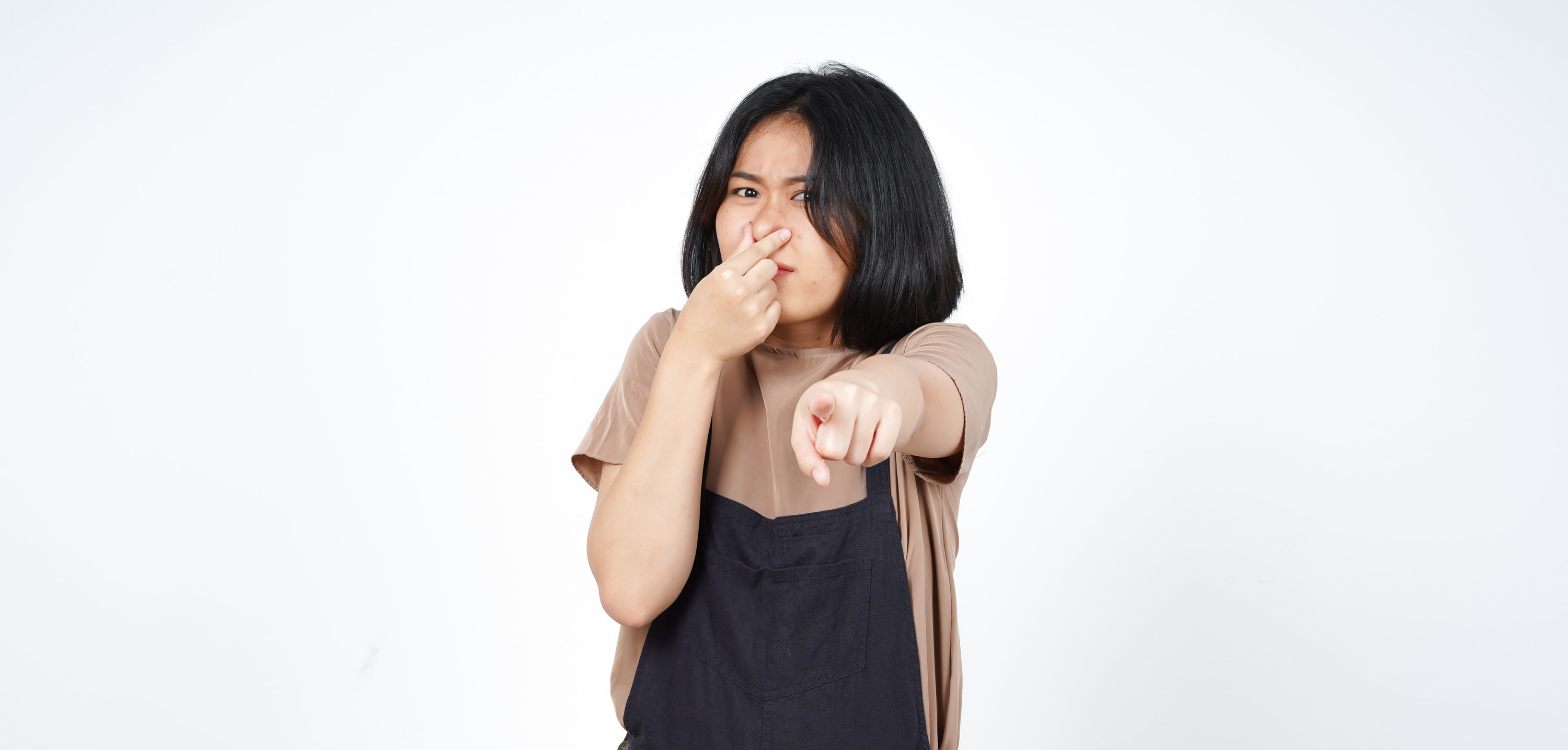 An image of a woman with short black hair in a bob, pointing at the camera with her nose held in her other hand as though she&#x27;s smelled something bad