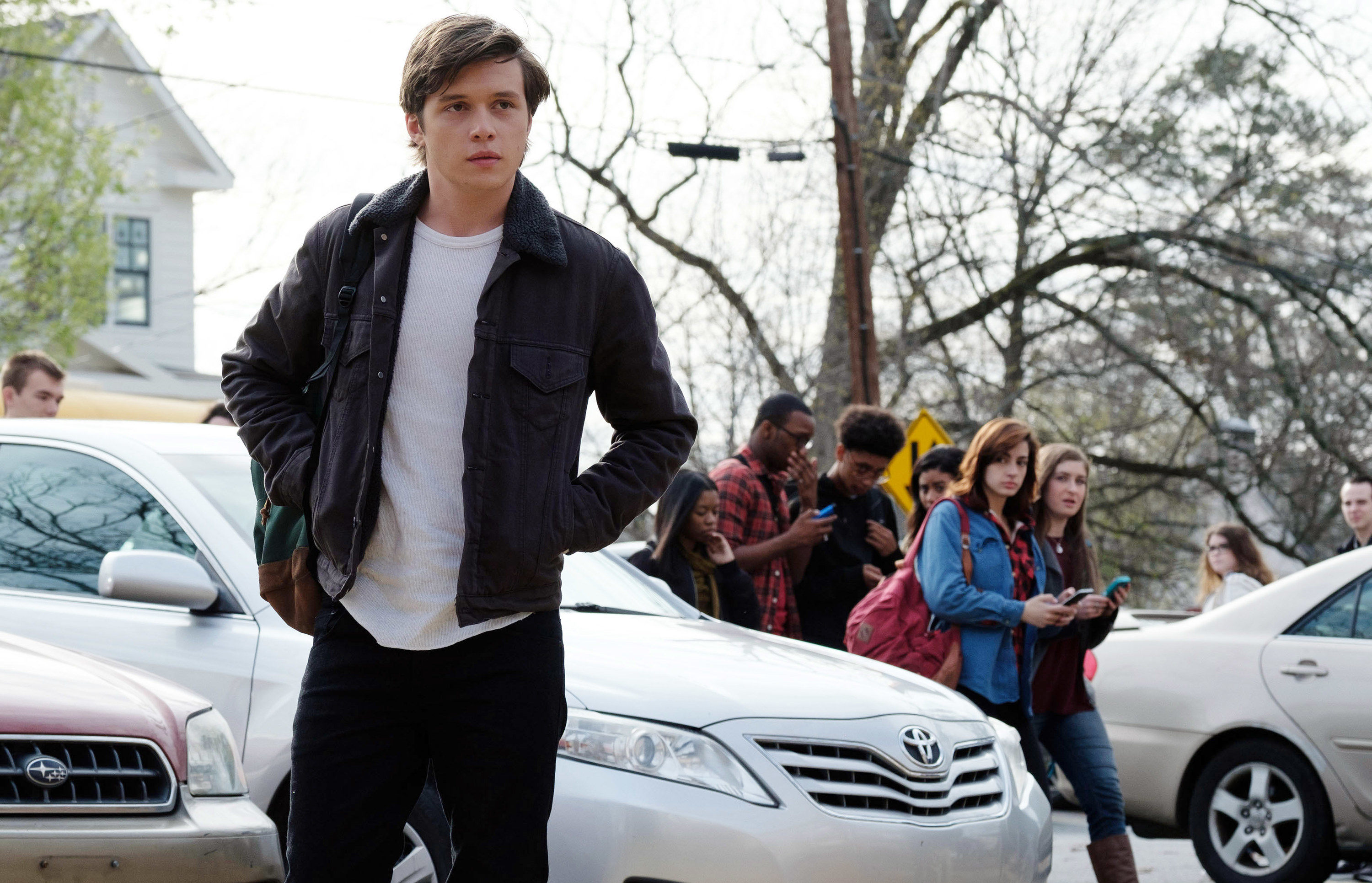 Young man stands by himself next to a car with other young people behind him