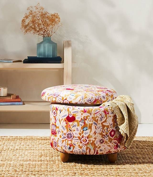 The storage ottoman with a floral design, the lid slightly off, and a throw blanket sticking out