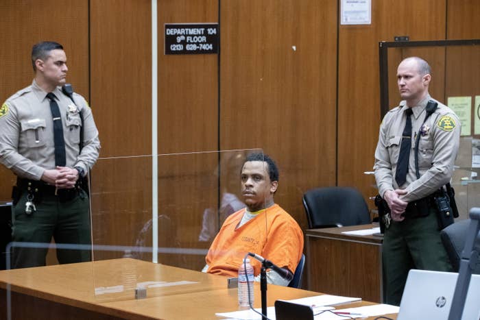 In a courtroom, two white police officers stand with their hands folded near a Black man wearing an orange jumpsuit seated at a table behind a transparent partition