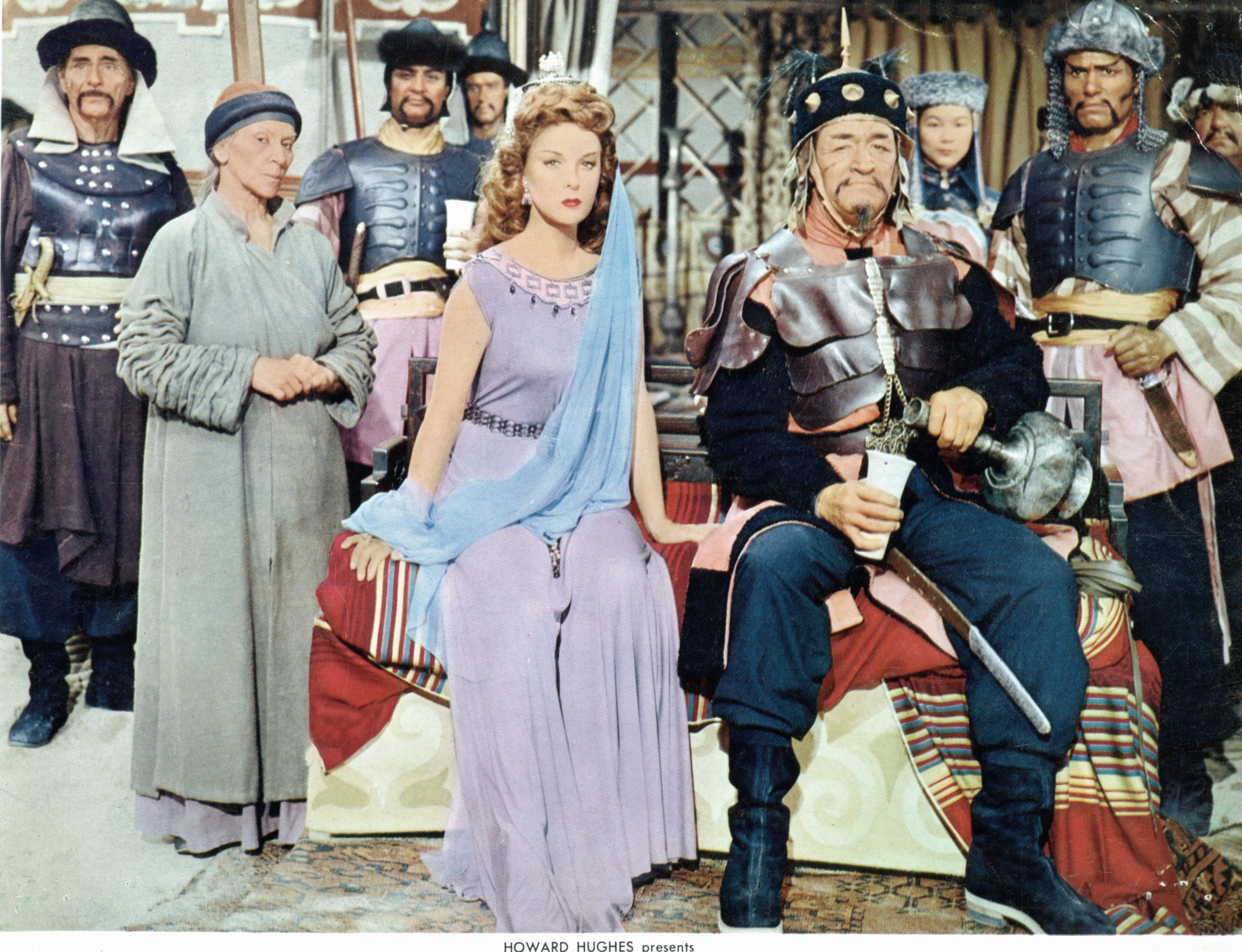 Susan Hayward and John Wayne sitting together surrounded by unidentified actors in a scene from the film &#x27;The Conqueror&#x27;
