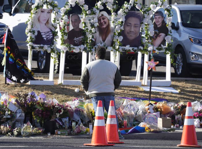 A person with their back to the camera faces five portraits of different people, lined by white roses, with dozens of bouquets of flowers on the ground