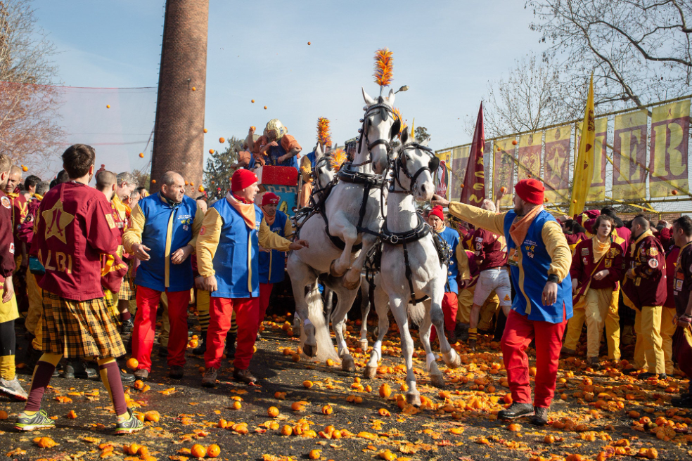 two white horses jump around surrounded by people in colorful costumes. on the ground, mashed oranges are everywhere