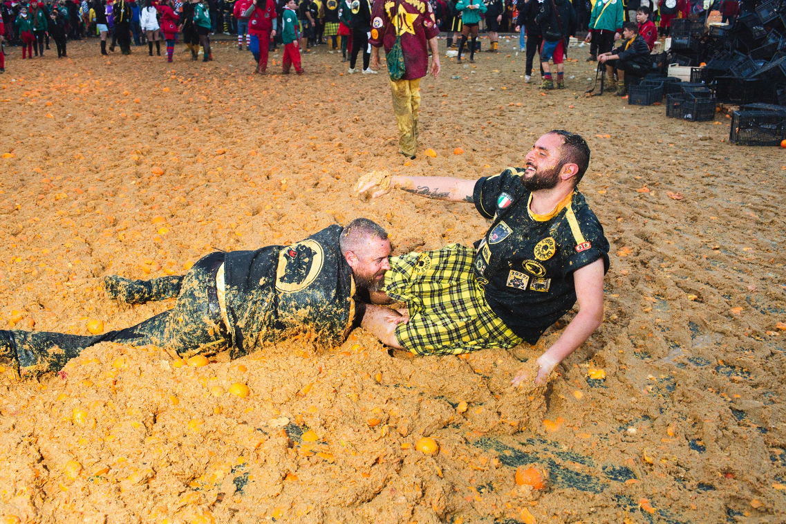 Two men writh around in a mass of orange pulp at the battle of the oranges. mashed oranges cover the entire floor as far as the eye can see.
