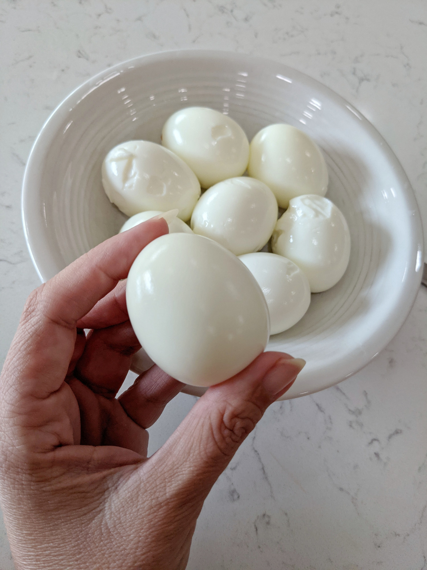 Woman&#x27;s hand holding a peeled Hard-boiled egg in front of bowl of eggs.
