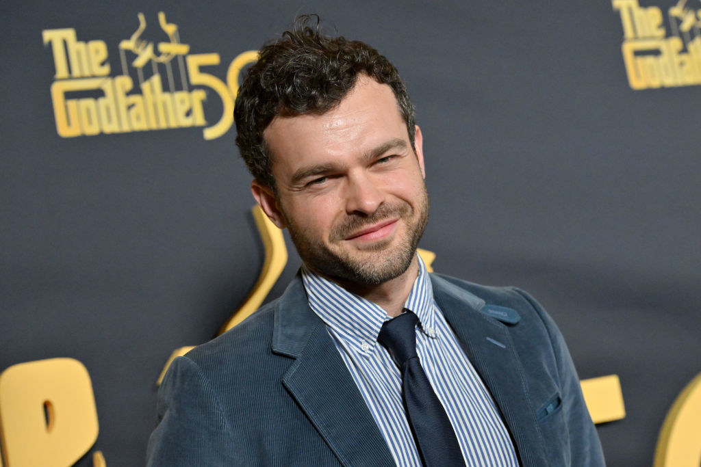 alden ehrenreich smiling in front of a the godfather poster on the red carpet