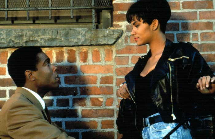 Black man is on his knee in suit while Black woman in a leather jacket sares at him intently