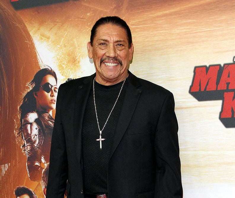 danny trejo smiling in front of a machete kills poster on the red carpet