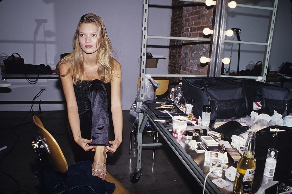 kate moss in the 90s adjusting her trousers while sitting in front of a vanity
