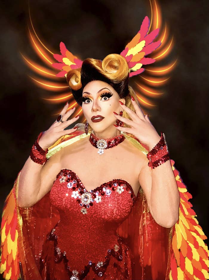 A drag queen wearing a strapless, bedazzled red dress holding up their hands with red nail polish to their face and surrounded by decorative red, orange, and yellow stripes