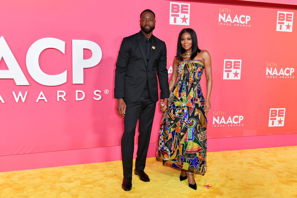 Dwyane in a monochromatic suit holding hands with Gabrielle, who&#x27;s wearing a multicolored, strapless geometric-print gown