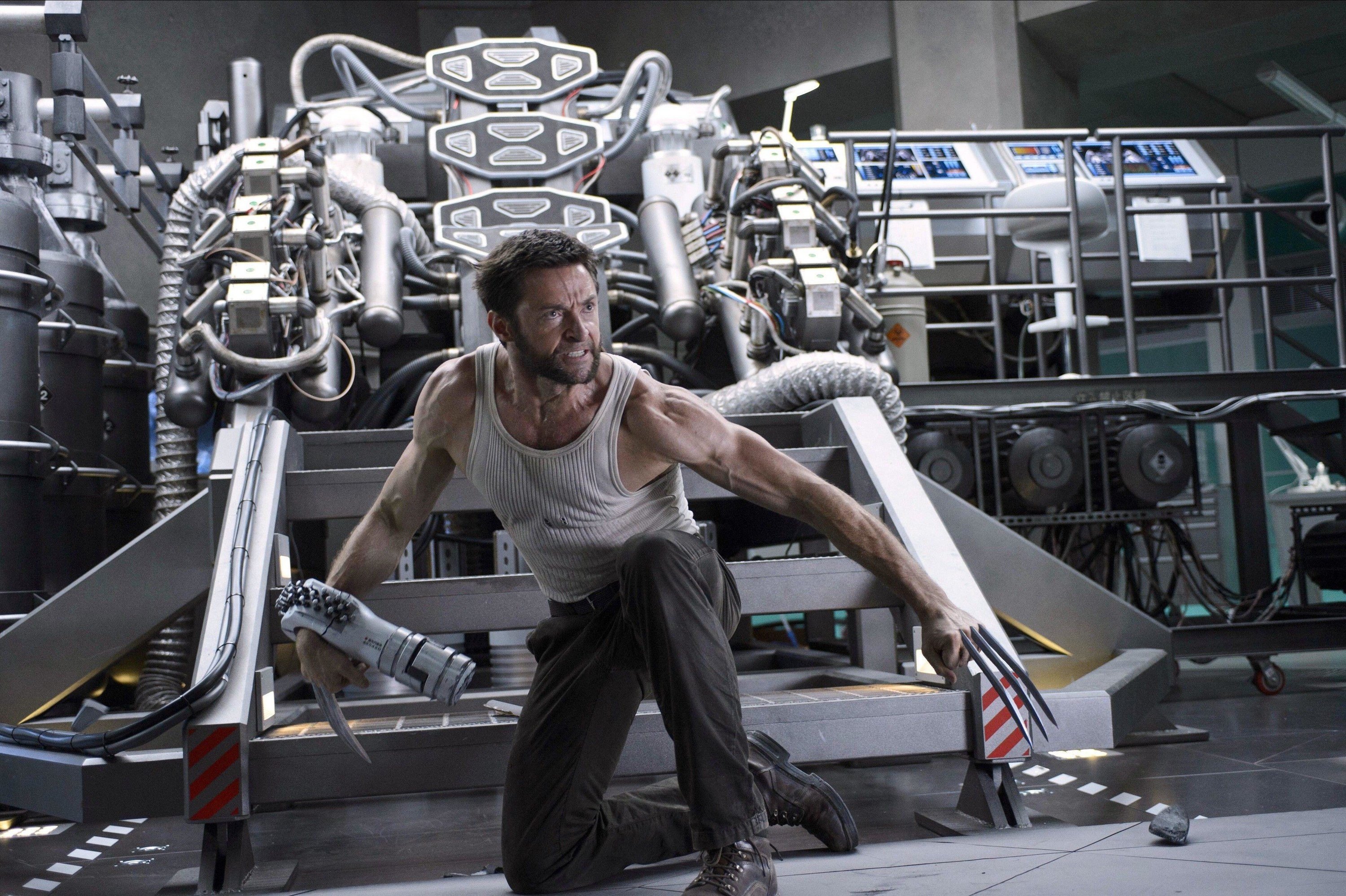 A man in a tank top with metal claws kneels near a giant machine