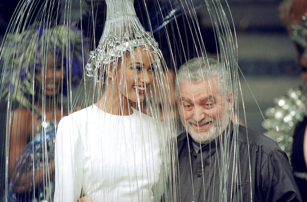 Paco Rabanne stands next to a tall white model wearing a white dress and a jellyfish-like hat on her head, with its long tentacles draping over the two of them