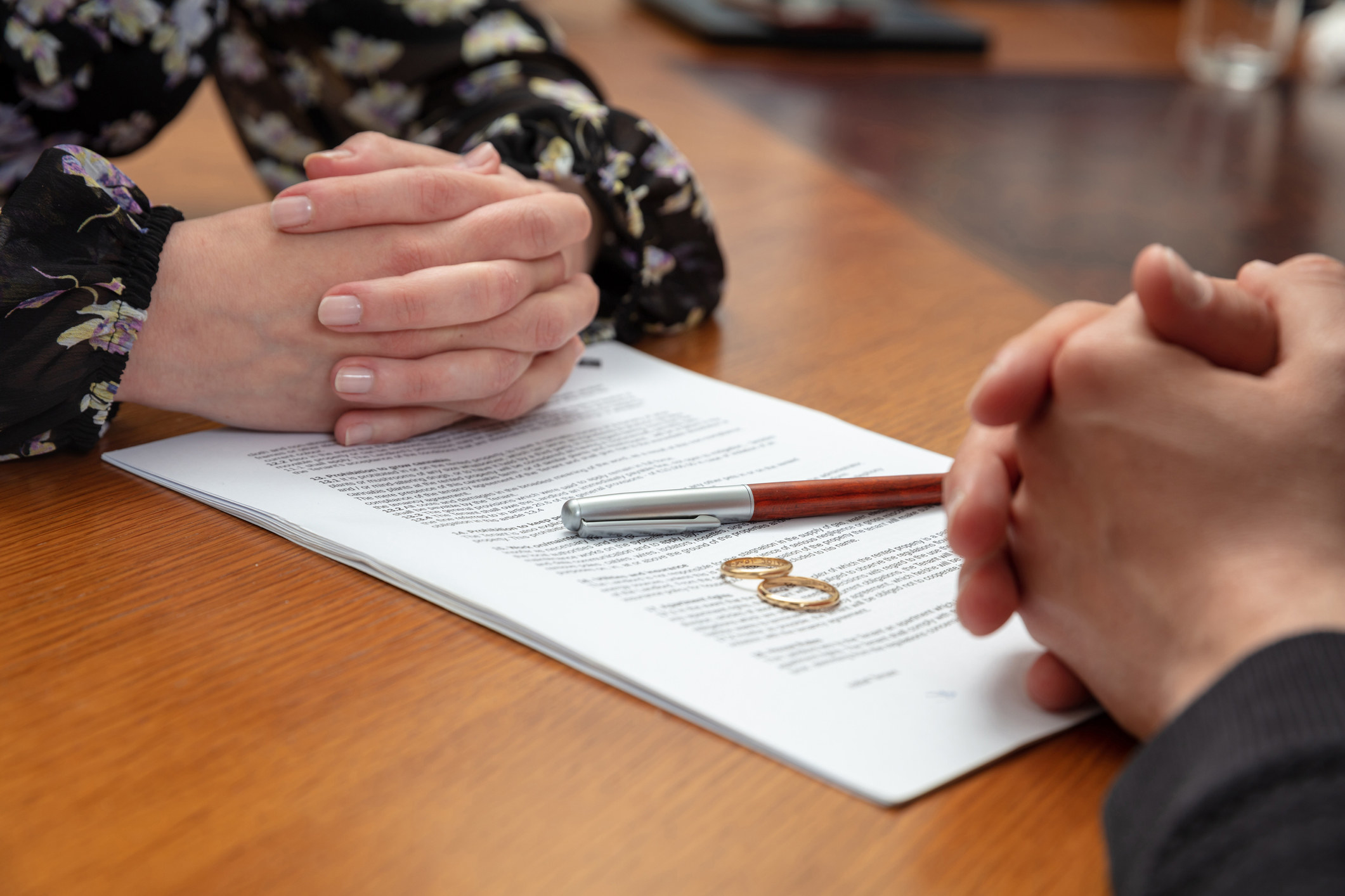 Two people sitting across from each other at a table with a divorce agreement, two rings, and a pen on it