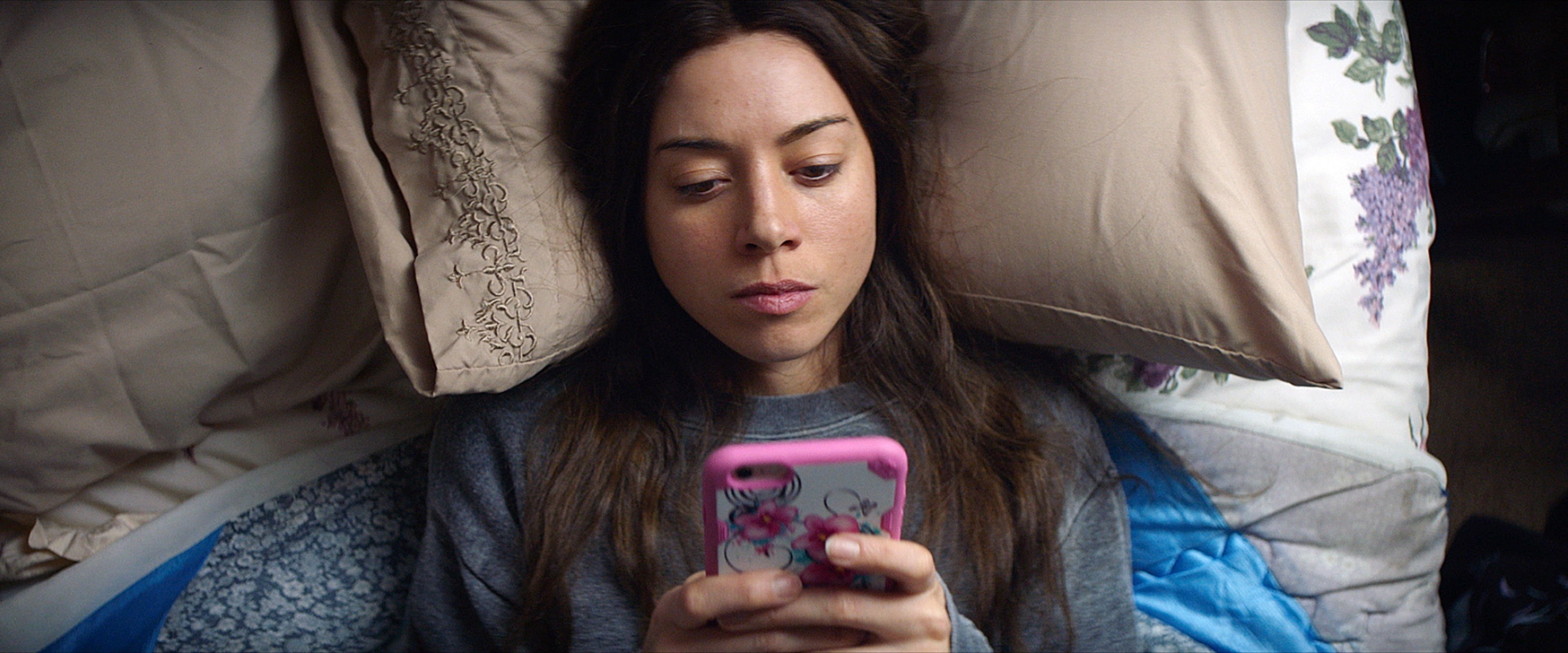 A woman lying in bed looking at her phone and looking glum