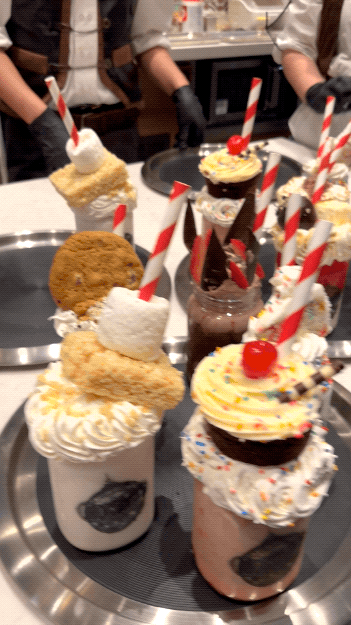 A dozen milkshakes in large mason jars with different flavors and toppings like cake, pie, and cookies