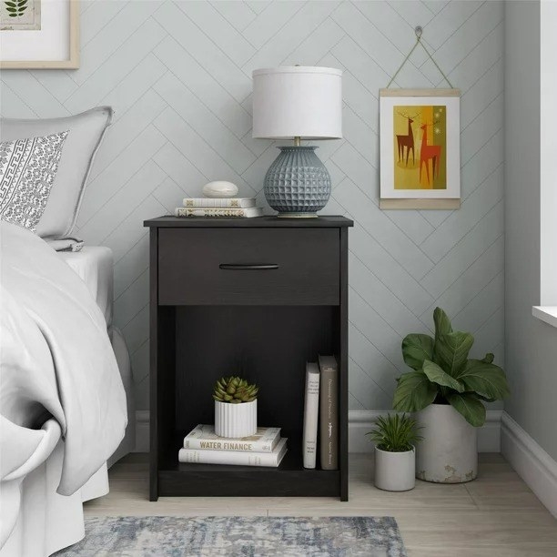 A black oak nightstand in a bedroom with a lamp resting on top and books stored inside