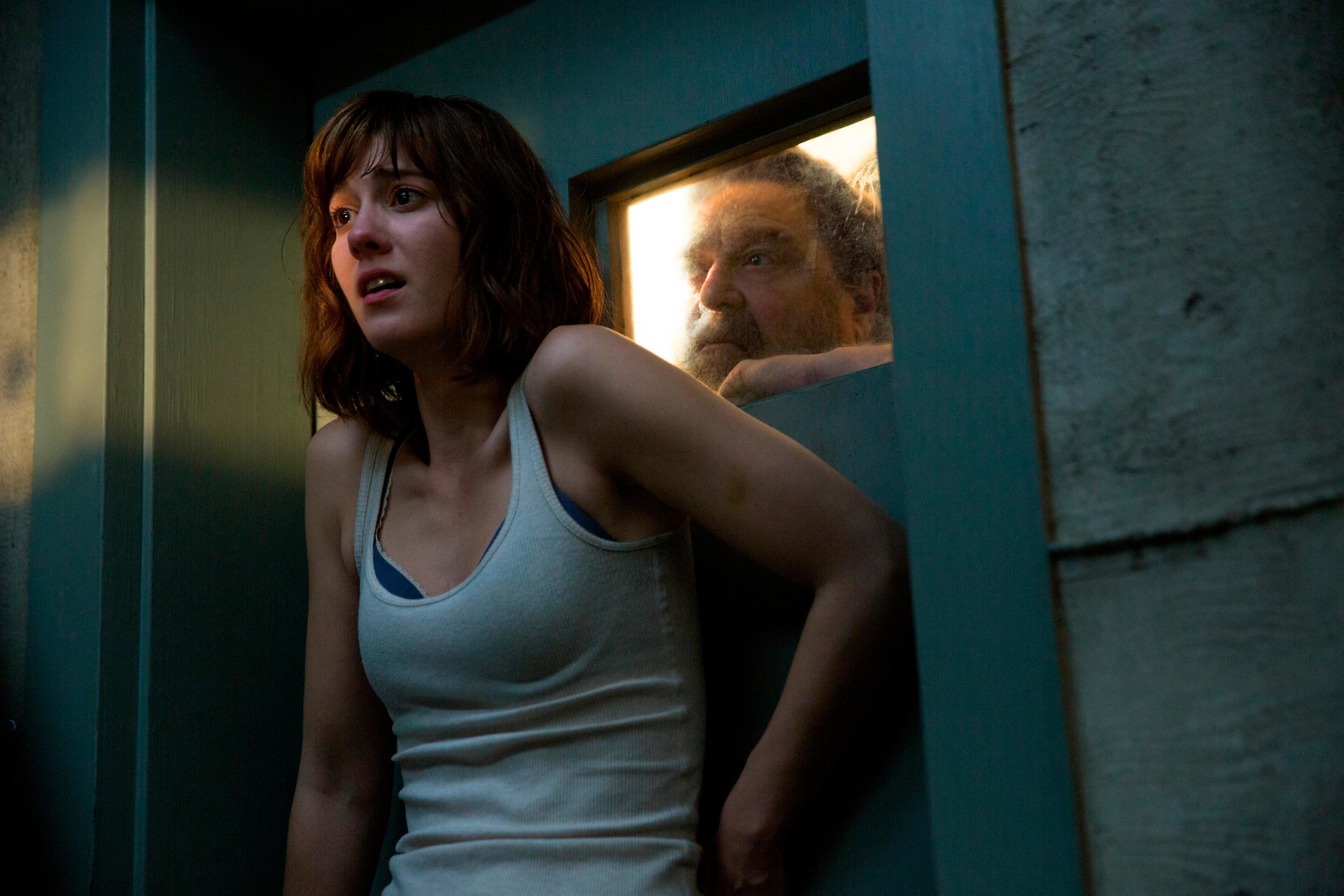 A terrified woman stares out of a vestibule with a man behind a steel door peering over her shoulder