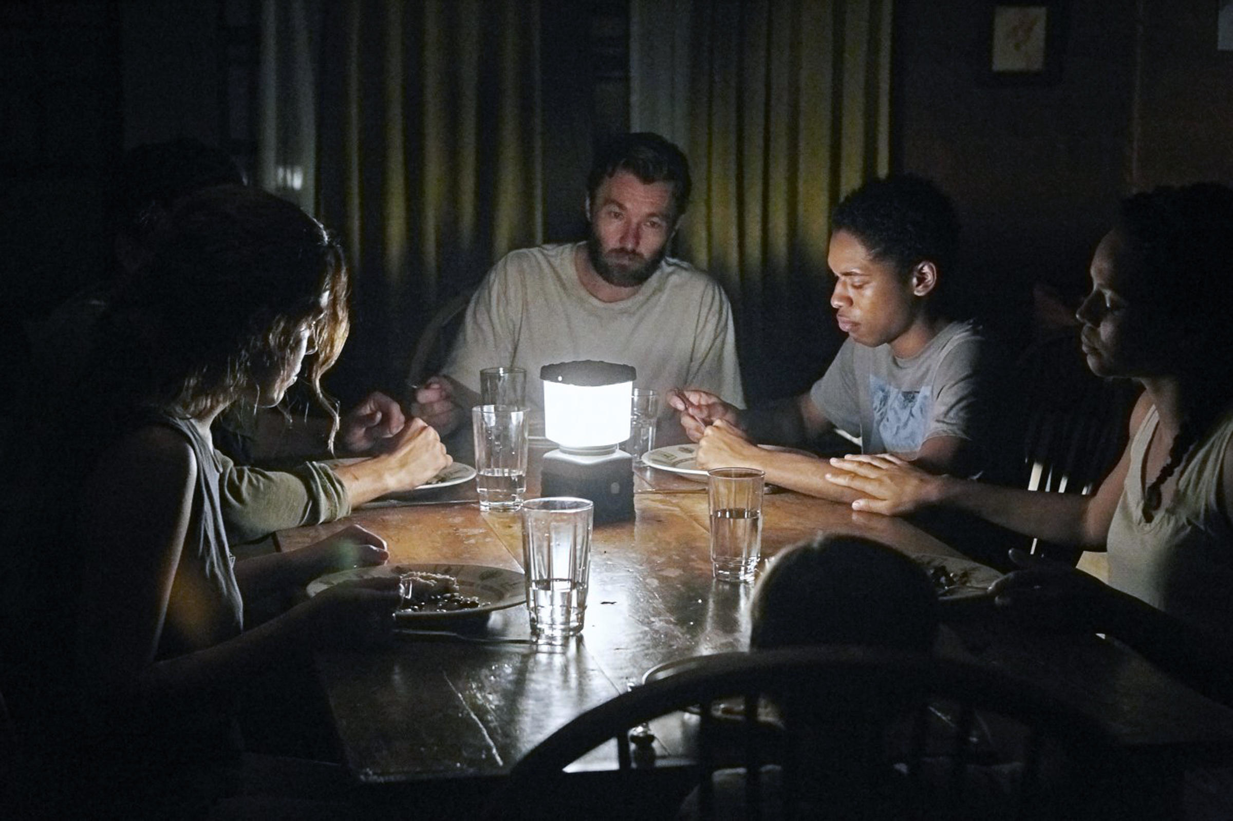A makeshift family gathers around for dinner in a dimly lit room illuminated by a lantern