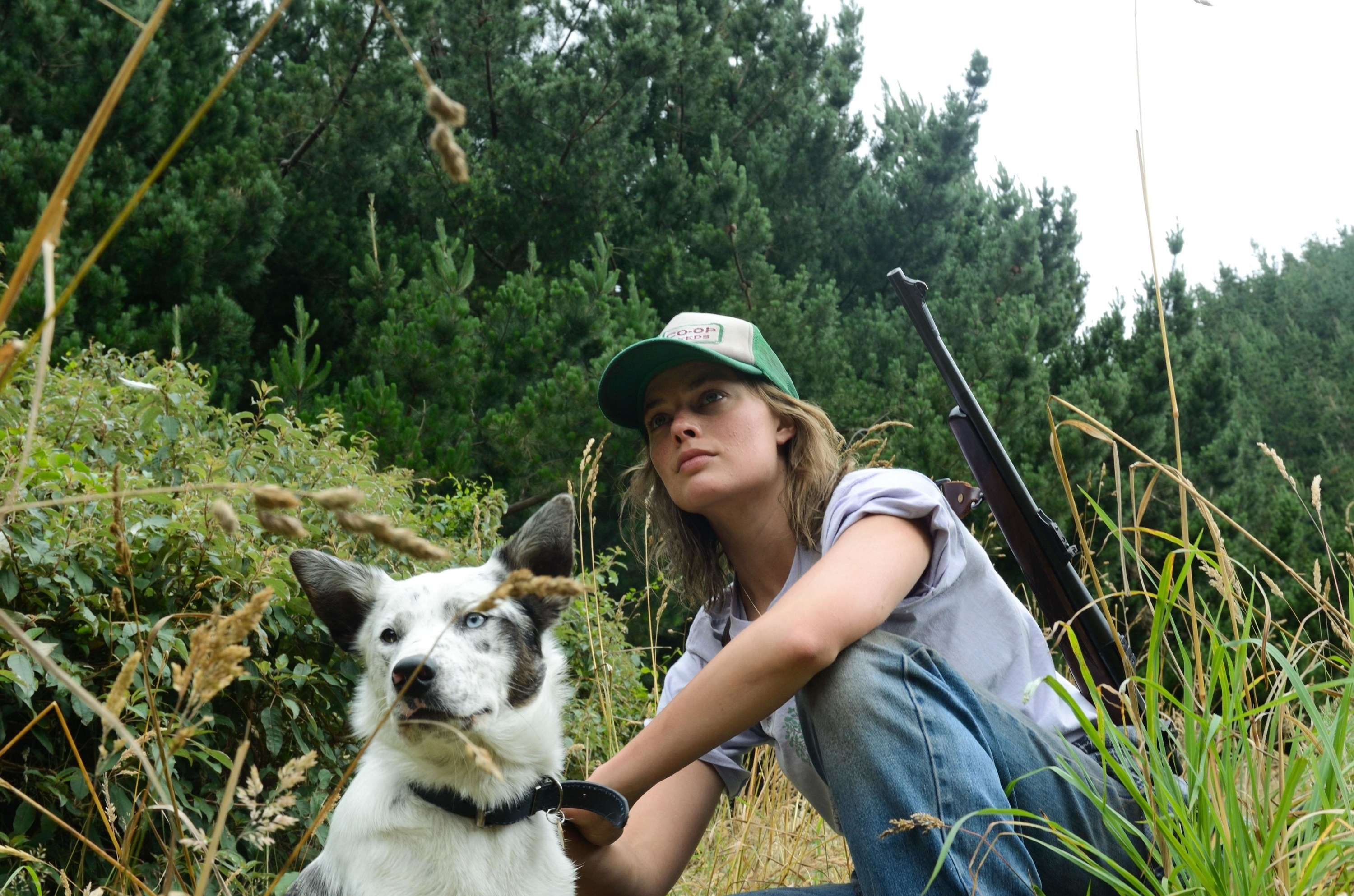 A woman with a rifle kneels near a bush with her spotted dog