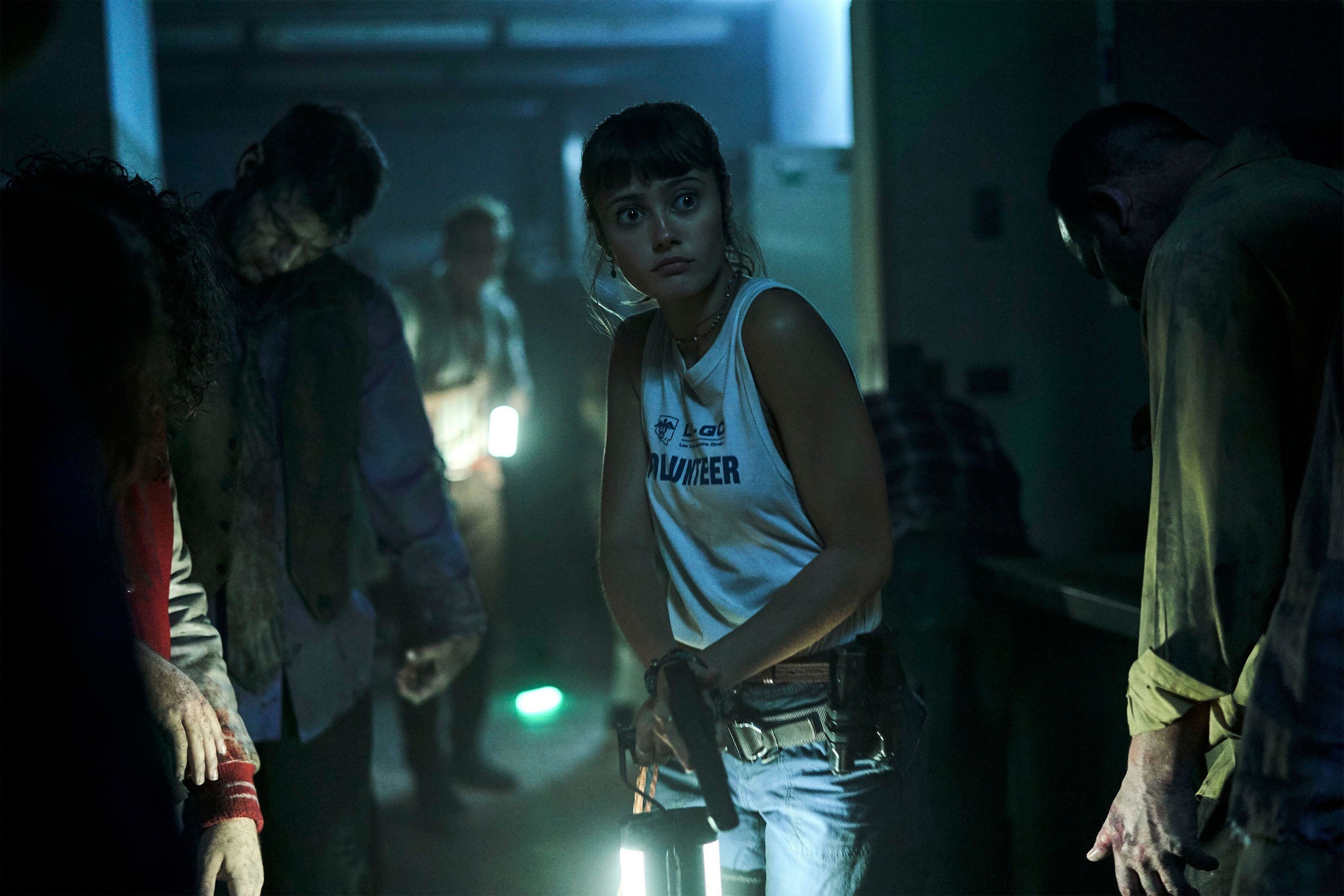 A young woman with a gun walks carefully through a hallway of zombies