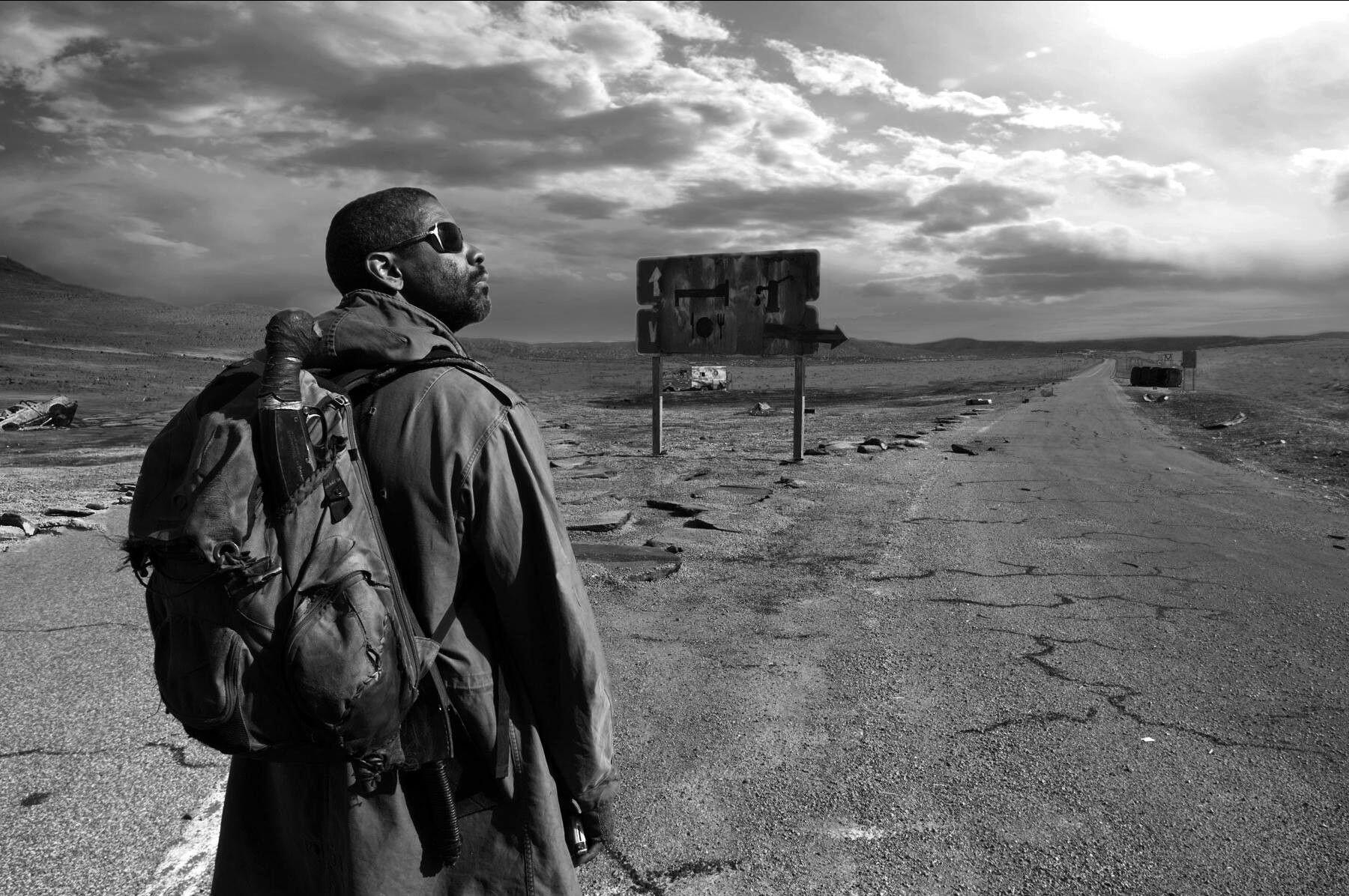 A man in a dirty coat with sunglasses walks down a long, colorless road