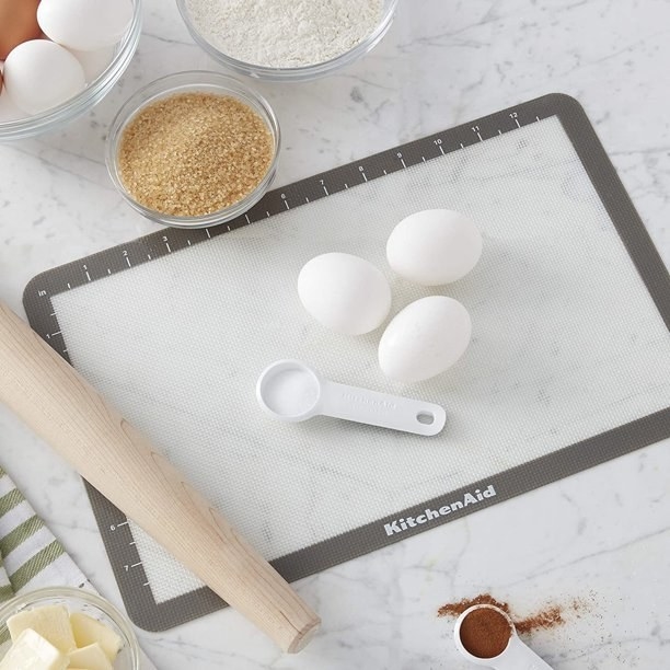 a white silicone baking sheet with a rolling pin, eggs, and a measuring coup on it