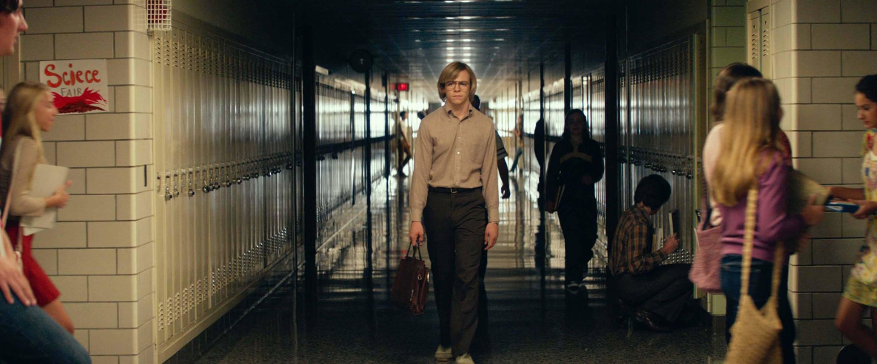 A young bespectacled man wanders through a dark high school hallway